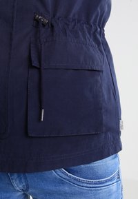 Granatowa kurtka ze sznurkiem w talii, z przednią kieszenią i gładką fakturą. Obecne kontrastowe szwy i metka z logo.