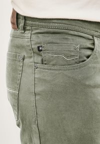 Grüne Baumwollhosen mit sichtbaren Nähten, einer kleinen Fronttasche und einem braunen Lederschild am Bund.