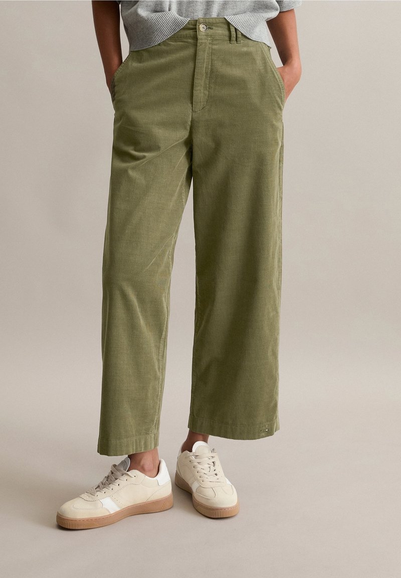 Personne portant un pantalon large vert olive et des baskets beiges, debout les mains dans les poches devant un fond uni.