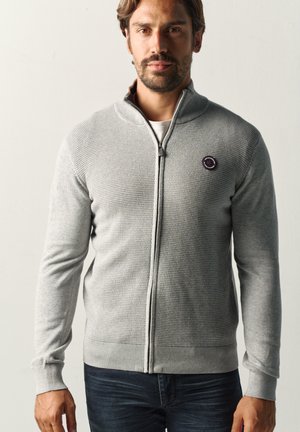 Mann trägt einen hellgrauen, strukturierten Zip-up-Pullover mit kleinem rundem Logo auf der linken Brust, kombiniert mit dunkelblauen Jeans.