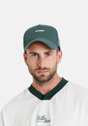 Groene baseballpet met een gebogen klep en witte logotekst. Wordt gedragen door een persoon in een wit shirt met donkergroene accenten.