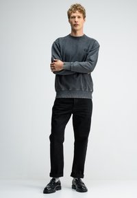 Grijze sweatshirt met een ronde hals en klein logo, gecombineerd met zwarte tapered broek en zwarte instappers, tegen een lichte achtergrond.