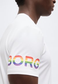 Άτομο που φοράει λευκό πουκάμισο με γράμματα στο μανίκι σε χρώματα του ουράνιου τόξου και κουκκίδες που σχηματίζουν τη λέξη "BORG" σε απλό φόντο.