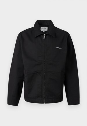 Musta vetoketjullinen takki, jossa on kaulus, pitkät hihat ja valkoinen "Carhartt"-logo rinnassa, esitetty yksinkertaisen taustan edessä.