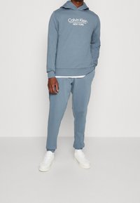 Felpa leggera blu con logo bianco "Calvin Klein NEW YORK", abbinata a pantaloni tapered coordinati e sneakers bianche. Tessuto morbido in misto cotone.