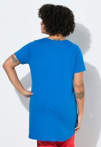 Personne aux cheveux bouclés portant une chemise à manches courtes bleue et un pantalon rouge, vue de dos, montrant des tatouages sur le bras gauche et le cou.
