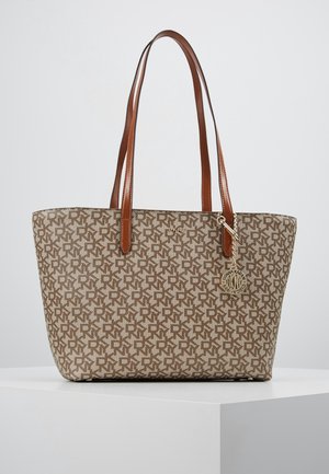 Brązowa torba typu tote z monogramem, podwójnymi skórzanymi uchwytami oraz okrągłą metalową zawieszką z napisem i logo marki.