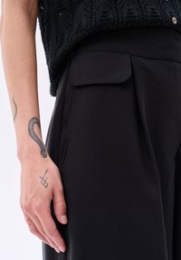 Zwarte, wijde broek met een platte tailleband en een zakdetail. Glad stoffen textuur, met een klein label aan de zijkant.