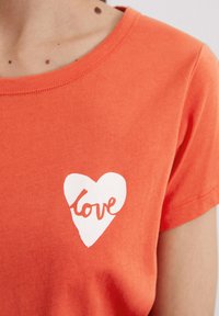Short-sleeve, round-neck t-shirt in bright orange cotton, featuring a white heart graphic with the word "love" in cursive.