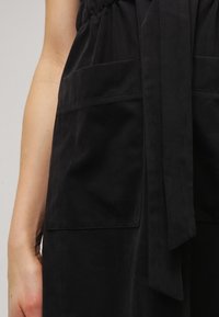 Robe noire avec un corsage ajusté doté d'une ceinture élastique, un détail noué et deux poches avant. Tissu lisse et mat avec une texture douce.