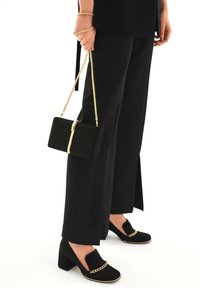 Clutch in velluto nero con catena dorata, abbinato a pantaloni larghi neri con spacchi laterali e scarpe con tacco blocco nere con dettagli dorati.