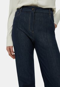 Pantaloni in denim blu scuro con una texture liscia, dotati di chiusura frontale con bottone, cuciture sottili e design classico a cinque tasche.