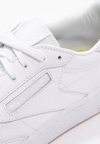 Vit sneaker med ovandel i slätt läder, texturerade sidopaneler, platta vita snörningar och gummisula. Har subtila märken på tungan.