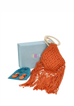 Bolso naranja de macramé con asas de anillas de madera y largos flecos, expuesto junto a unos pendientes a juego sobre una tela verde azulado y una caja azul claro.