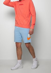 Hoodie corallo con testo "JUST DO IT", abbinato a pantaloni corti blu chiaro con un grande logo arancione Nike. Sneakers bianche completano l'outfit.