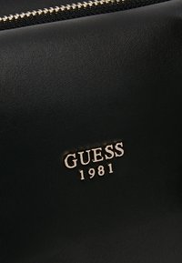 Zwart synthetisch materiaal met een gladde textuur, met een gouden logo waarop "GUESS 1981" staat. Ritsdetails zichtbaar.