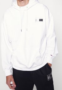 Felpa bianca con cappuccio, tasca frontale, patch logo argento, cordini e polsini a costine. Indossata con pantaloni della tuta neri con logo.