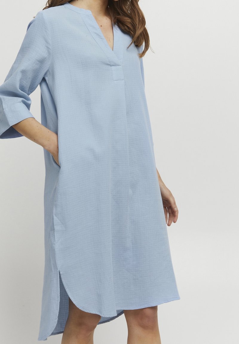 Femme portant une robe ample bleu clair, longueur genou, avec des manches trois-quarts et des poches latérales, debout devant un fond clair uni.