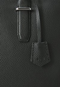 Detaliu al unei genți negre din piele, cu o suprafață texturată, accesorii din metal argintiu și un etichetă din piele cusută, embosată cu "BOSS."