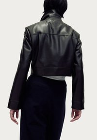 Veste en cuir noir recadrée avec un col montant et des détails de couture visibles, associée à un pantalon foncé. Texture lisse et coupe ajustée.