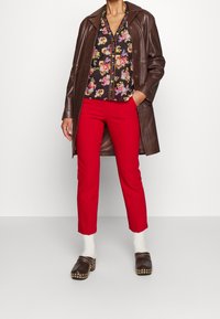 Manteau en cuir marron, blouse florale avec un fond noir et des motifs colorés, pantalon slim rouge, et sabots noirs avec des ornements en studs.