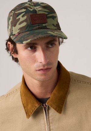 Mand iført en grøn og brun camouflagekasket med en brun lap og en beige jakke med en brun kordfløjls-krave, kiggende til siden.