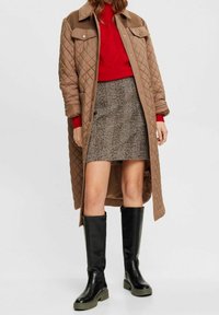 Žena v dlouhém prošívaném béžovém kabátu, červeném svetru, hnědé kostkované sukni a černých vysokých botách, stojící na jednolitém pozadí.
