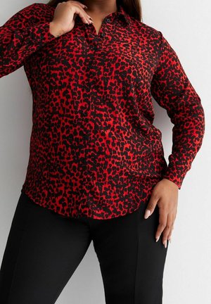 Frau trägt ein langärmeliges, rot-schwarz leopardenmuster Knopfhemd mit schwarzen Hosen, posiert mit einer Hand am Kragen und einer an der Hüfte.