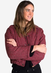 Chaqueta recortada a cuadros rojos y negros con una tela texturizada, botones a presión y un diseño estructurado, llevada sobre una blusa negra.