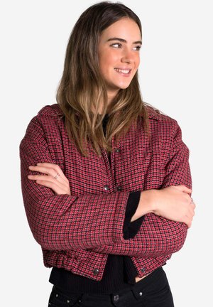 Veste courte à carreaux rouge et noir avec un tissu texturé, boutons-pression et un design structuré, portée sur un haut noir.