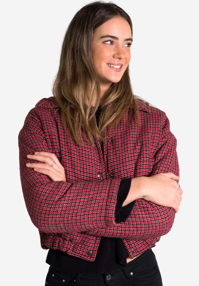 Chaqueta recortada a cuadros rojos y negros con una tela texturizada, botones a presión y un diseño estructurado, llevada sobre una blusa negra.
