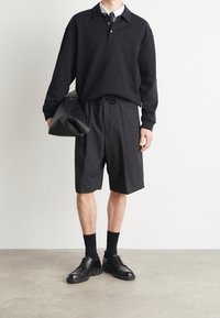 Musta pitkähihainen poolopaita valkoisen paidan päällä, yhdistettynä mustiin shortseihin ja mustiin nahkakenkiin, kädessään musta laukku.