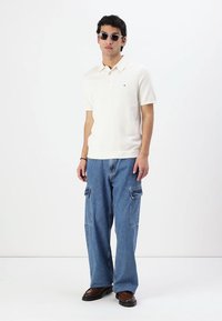 Λευκό πόλο μπλουζάκι από βαμβάκι, συνδυασμένο με ανοιχτό μπλε cargo τζιν, που διαθέτουν μεγάλες πλαϊνές τσέπες, και καφέ παπούτσια. Τα αξεσουάρ περιλαμβάνουν σκούρα γυαλιά ηλίου.