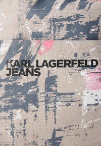 Μπεζ ύφασμα με αφηρημένα γκρι και ροζ σχέδια· διαθέτει το κείμενο "KARL LAGERFELD JEANS" με έντονα μαύρα γράμματα· υφή με ανάγλυφη επιφάνεια.