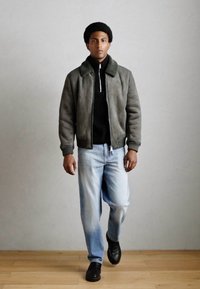 Sivi suede bomber jakna s tamnozelenim ovratnikom, crni zip-sweater ispod, svijetloplave traperice i crne cipele na drvenom podu.