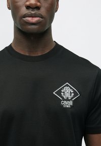 Musta puuvillainen t-paita, jossa on timantinmuotoinen valkoinen kirjailtu logo. Lyhyet hihat ja pyöreä kaula-aukko. Sileä pinta.