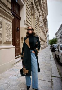 Długi czarny płaszcz z beżowymi akcentami i wzorem w panterkę, czarny golf, jasne szerokie dzwony jeansowe oraz czarna torba. Otoczona miejską architekturą.