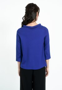Kék, háromnegyedes ujjú felső, cseppnyakú, sima textúrával, fekete nadrággal párosítva, amely egyszerű dizájnnal és letisztult vonalakkal rendelkezik.