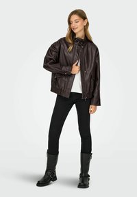 Giacca in finta pelle marrone con colletto alto, zip frontale, orlo inferiore elastico e tasche laterali, abbinata a jeans skinny neri e stivali neri.