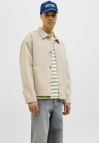 Beige denim jacket with gray buttons over a green and white striped shirt. The model wears light blue jeans and a blue cap with text.