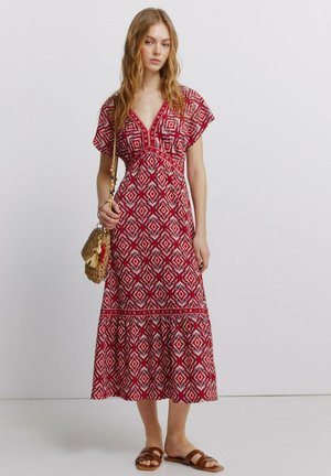 Kvinde iført en midi-kjole med rød og hvid geometrisk mønster, brune sandaler og bærende en flettet håndtaske med kvast-detaljer.