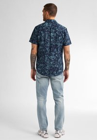Camicia a maniche corte, blu scuro, con un motivo di foglie tropicali in teal. Abbinata a jeans azzurri chiari e sneakers bianche, vista da dietro.