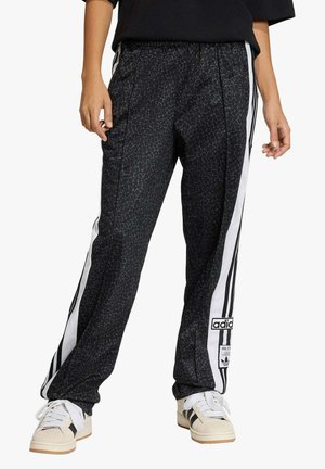 Persona che indossa pantaloni della tuta Adidas neri con motivo a fantasia e strisce laterali bianche, e sneakers beige con strisce nere.