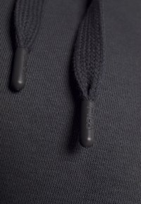 Primer plano de una sudadera negra con capucha, que presenta cordones de tejido trenzado con puntas de goma negra grabadas con el logotipo de Fila, sobre un fondo de tejido texturizado.