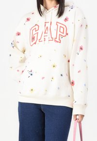 Bílá mikina s kapucí s květinovým vzorem, která má červené logo "GAP", klokaní kapsu a nastavitelné stahovací šňůrky u kapuce.