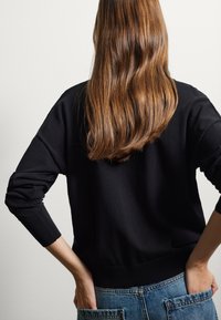 Schwarzer Pullover mit lockerer Passform, gerippten Bündchen und Saum. Kombiniert mit hellblauen Jeans mit aufgesetzten Taschen. Angenehme, glatte Textur.