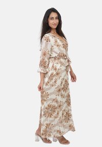 Robe longue crème avec des motifs floraux marron, décolleté en v et manches trois-quarts. Présente une ceinture assortie et une silhouette fluide. Talons transparents.