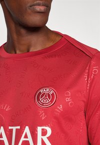 Punane särk ümmarguse kaelusega, millel on nähtav Paris Saint-Germaini logo ja peen reljeefne "Jordan" muster kogu kangas.