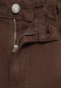 Tissu marron avec une fermeture éclair visible et un bouton, présentant un tissage texturé et des détails de couture. La fermeture éclair est en métal, positionnée à l'avant.