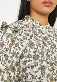 Blouse fleurie en tissu blanc avec des motifs de fleurs vertes et orange, dotée d'un col haut et de détails à volants sur les épaules.